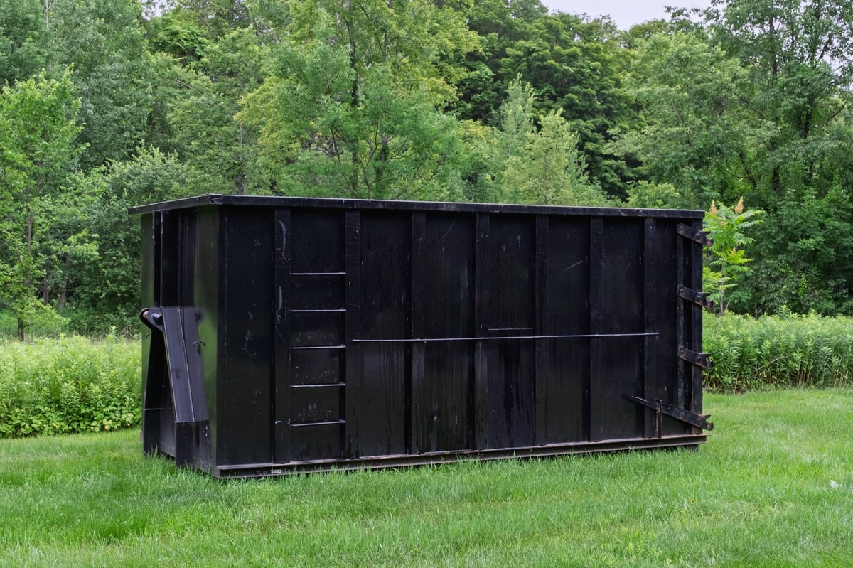 A large industrial black roll-off dumpster placed on a grassy lawn with lush green trees behind
