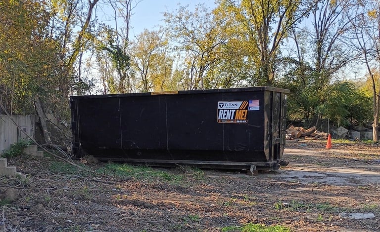 Black dumpster rental container on gravel ground with autumn trees in background
