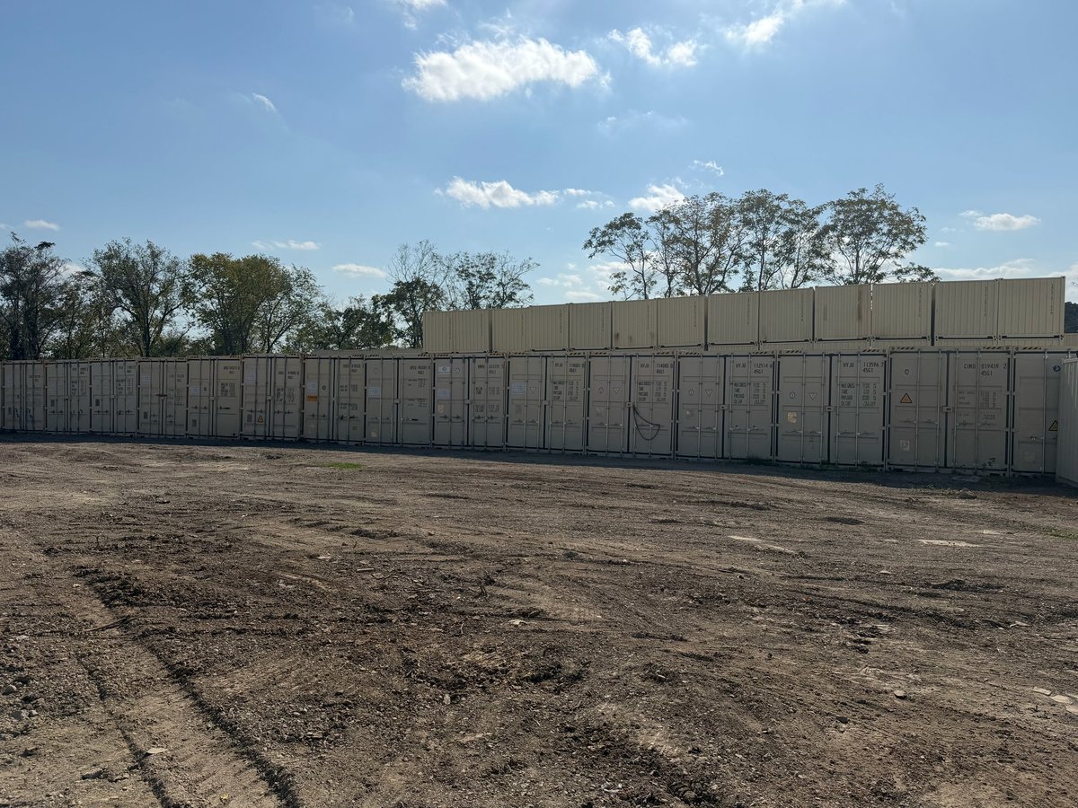 Empty dirt lot with rows of beige metal storage units or sheds under clear blue sky with trees in background