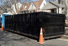 Large black dumpster with orange traffic cone in residential driveway with houses in background