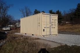 Beige metal shipping container positioned on gravel in an outdoor area with bare trees and residential buildings in background