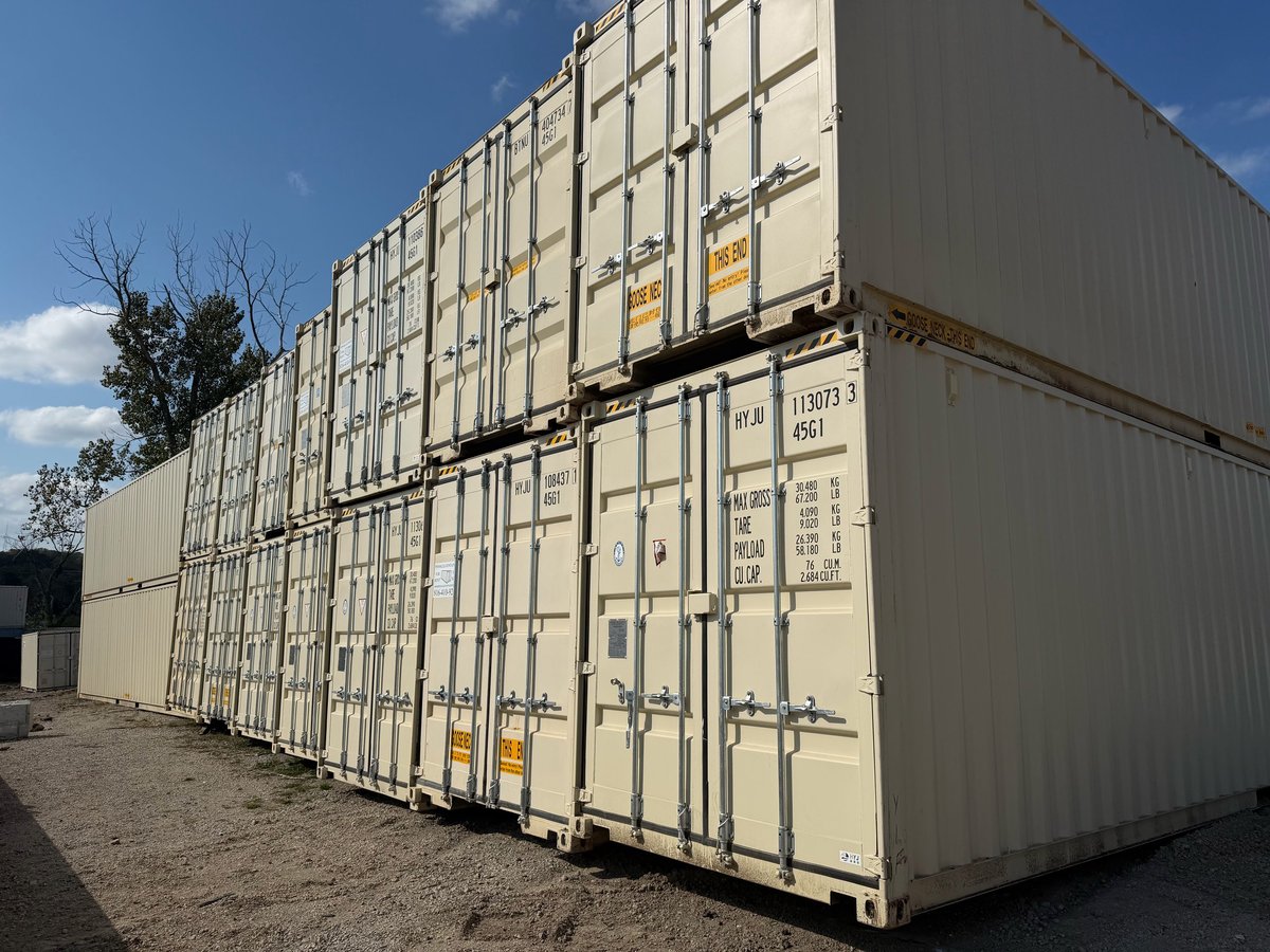 Stacked beige and gray shipping containers with metal doors and hinges against a blue sky.