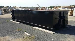 Black industrial dumpster on asphalt with industrial yard and buildings in background